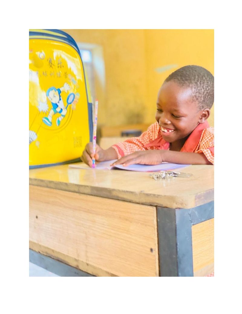 Ahmad at his school desk smiling broadly while writing in his notebook, yellow bag beside him