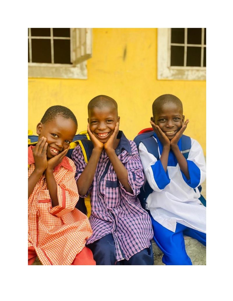 Three boys including Ali sitting together against a yellow wall, all smiling
