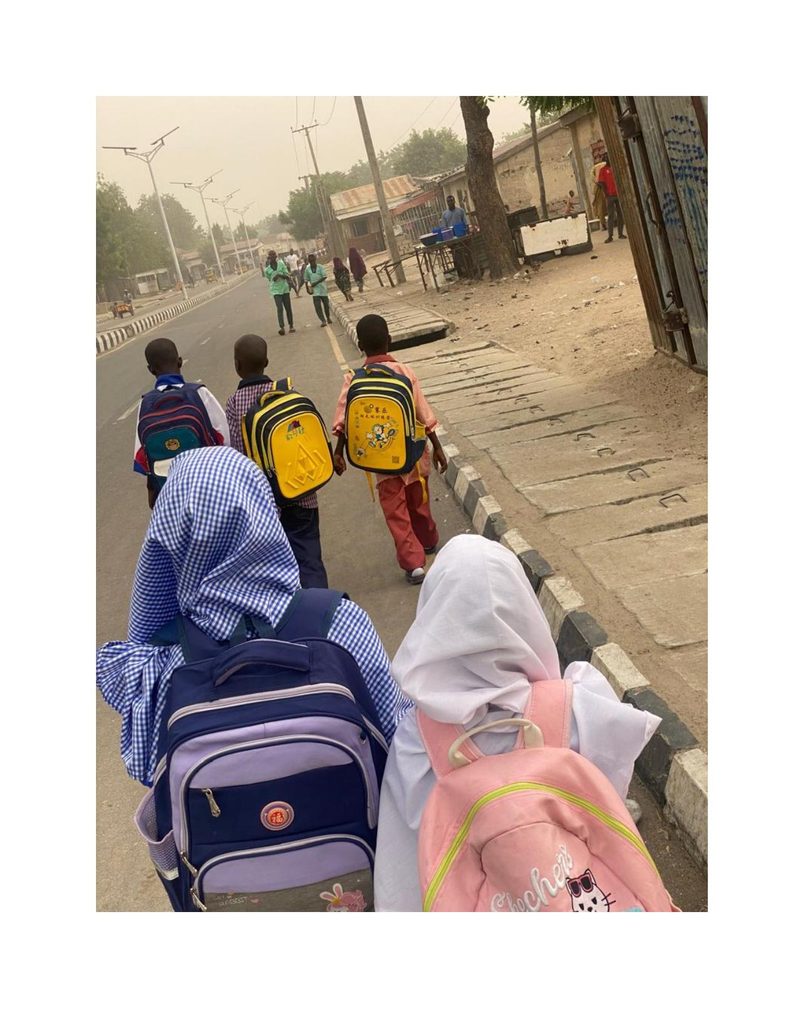 A group of children including Bilkisu walking along a dusty road to school carrying backpacks