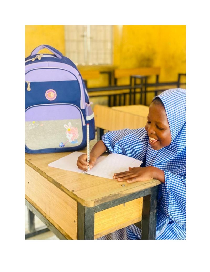 Bilkisu bent over her notebook writing with a pencil, smiling