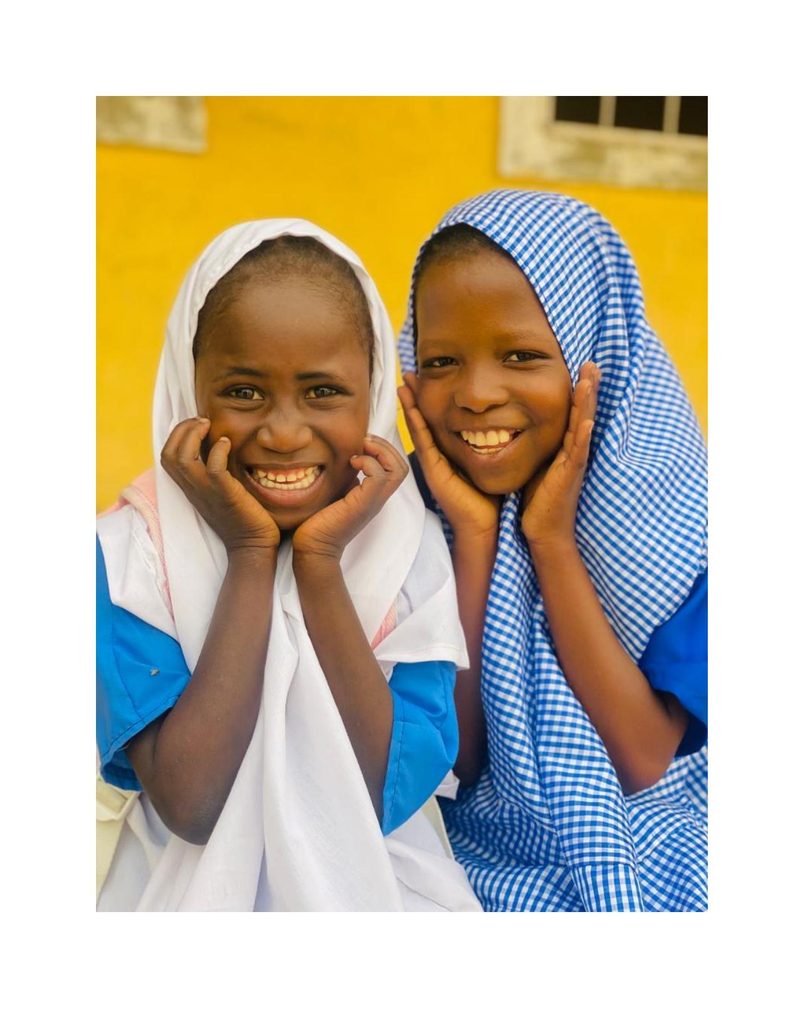 Bilkisu and her friend laughing joyfully together against a yellow wall