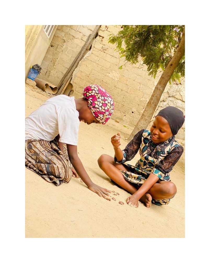 Fatima and a friend sitting cross-legged in the sand playing a seed game outdoors