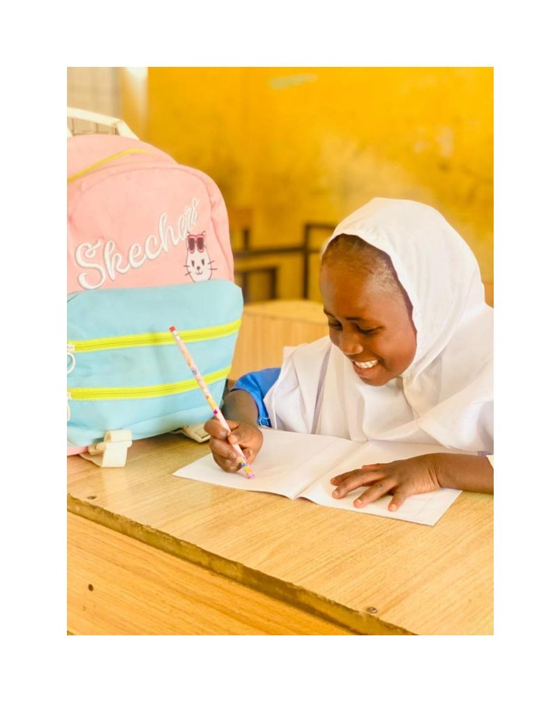 Fatima sitting at her school desk smiling while writing in her notebook, pink Skechers bag beside her