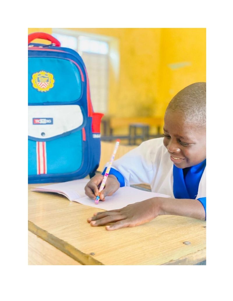 Musa bent over his notebook at his school desk, smiling as he writes, blue bag beside him